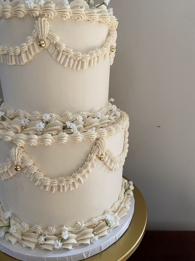 A two tier cake sports scallops of buttercream piping in the vintage lambeth style. There's also accents of gold pearls and tiny white flowers tucked into the borders.