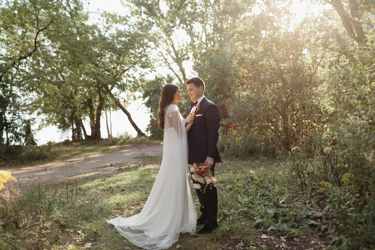 A bride and groom stand in a sunlit wooded area, captured beautifully by a talented Toronto photographer. The bride, in a flowing white dress holding flowers, gazes affectionately at her groom in his dark suit.