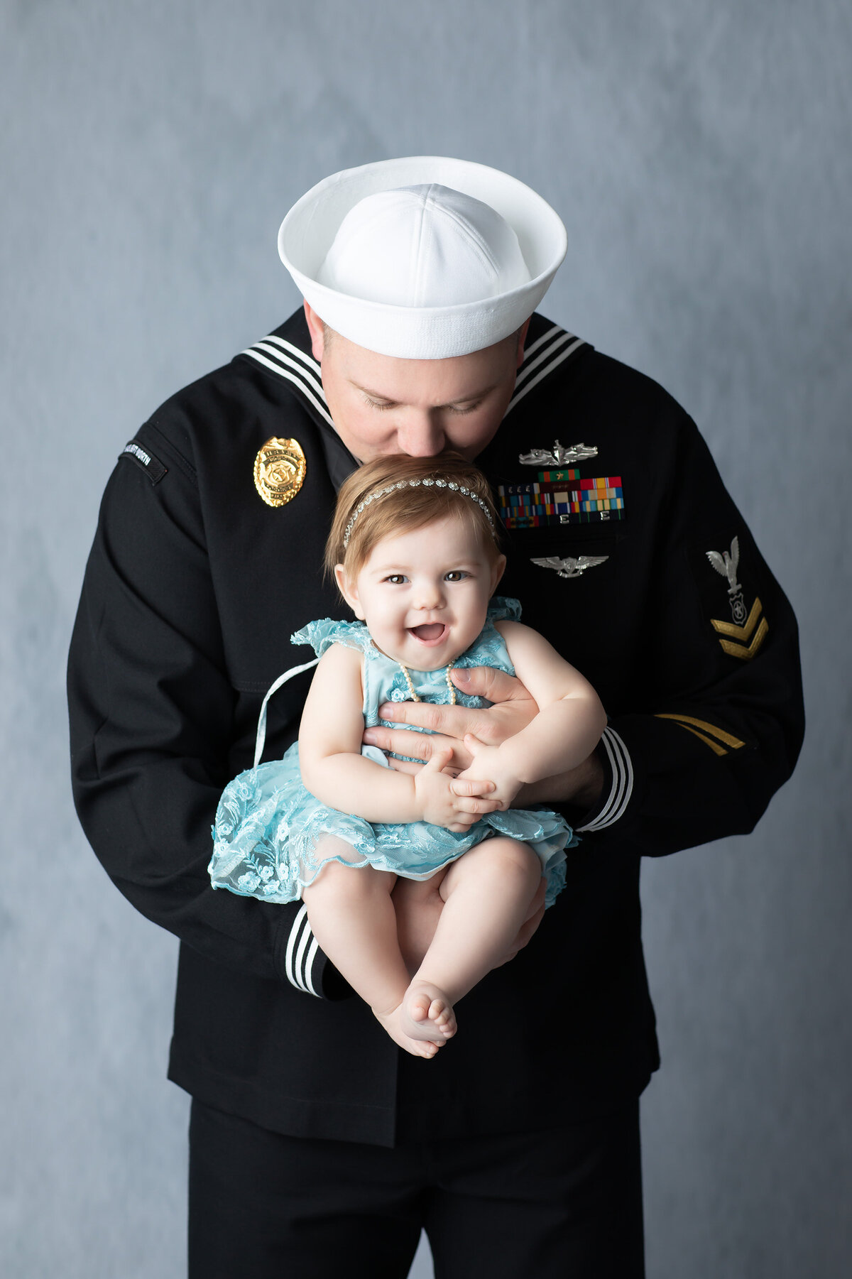 A proud Navy dad in uniform gently kisses his smiling baby girl on the head during a DFW milestone photography session.