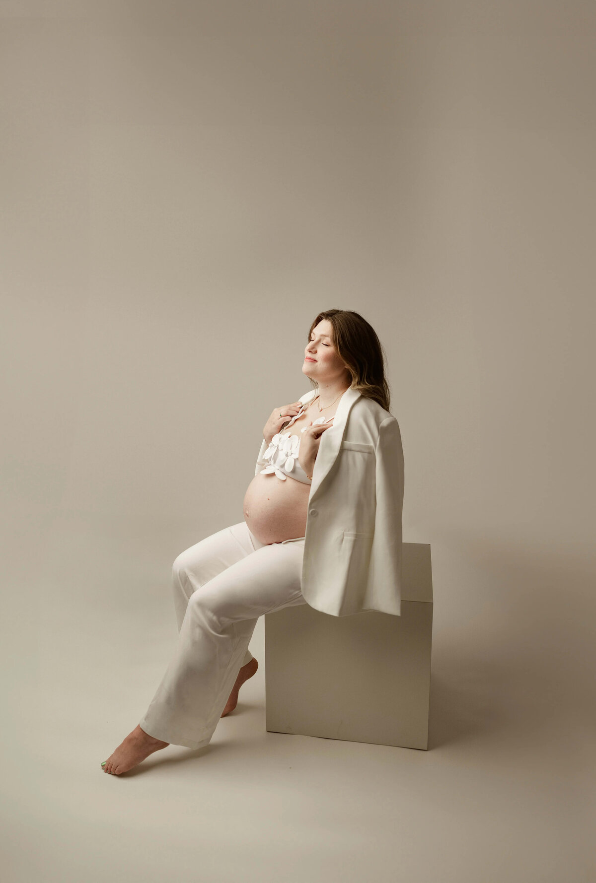 Seated maternity pose with white blazer and pants in minimalist studio 