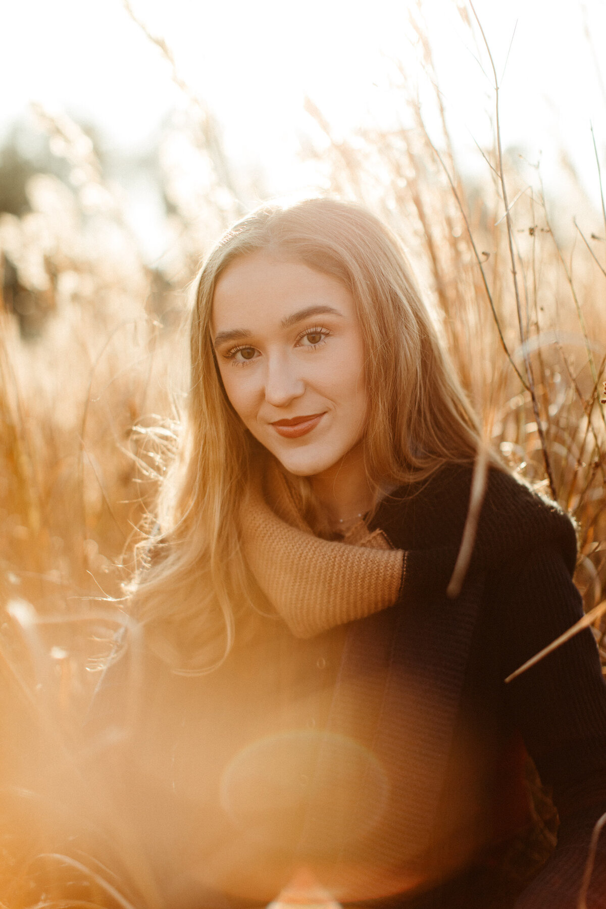 High school senior wearing a black sweater dress and scarf soft smiling in a field at sunset