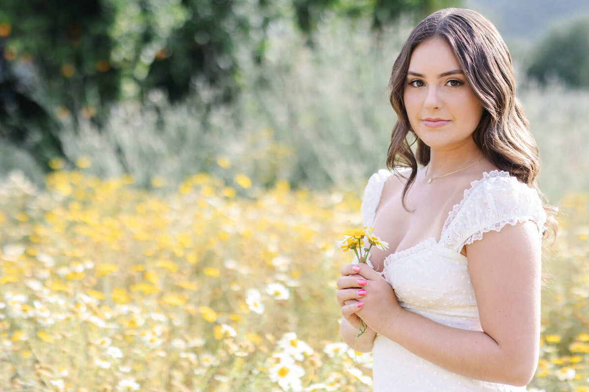 A woman in a white dress stands in a field of yellow and white flowers, holding a small bouquet. She has long brown hair and is looking at the camera, with a soft focus background of greenery.