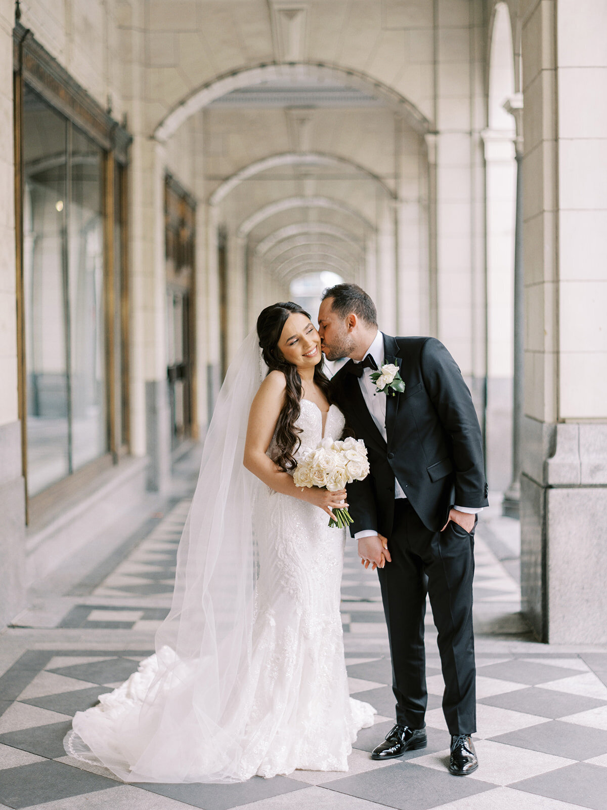 A bride and groom in wedding attire stand in a tiled hallway at the Fairmont Palliser Calgary. The groom kisses the bride on the temple as she smiles, holding a bouquet of flowers. They are standing under an arched ceiling, capturing a classic Calgary wedding moment.