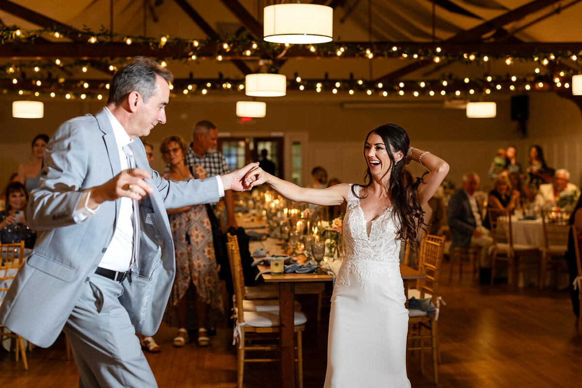 The bride dances with an older man in a light gray suit, both smiling broadly, in the warmly lit reception hall.