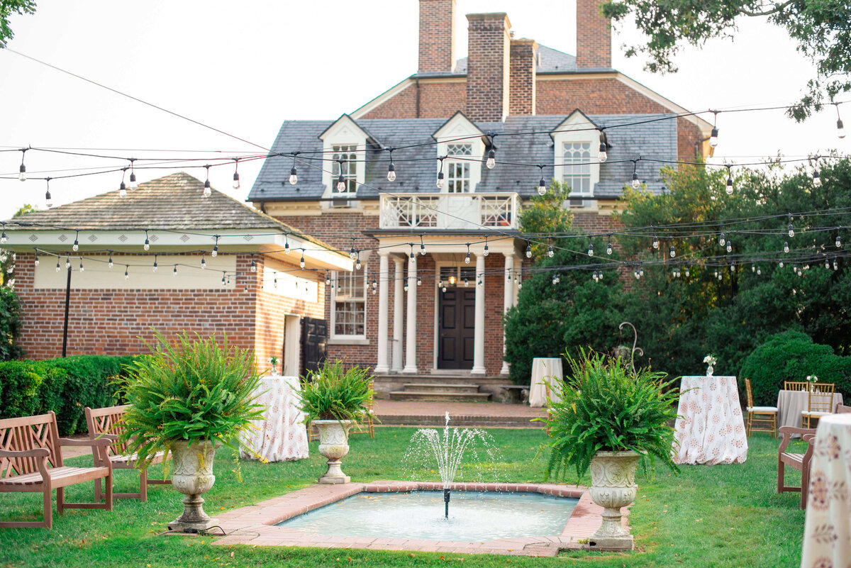 A brick estate with a columned entrance stands behind a small fountain surrounded by lush ferns. String lights hang overhead, and tables with white cloths are set up on the green lawn. Benches are arranged nearby, creating an elegant outdoor setting.
