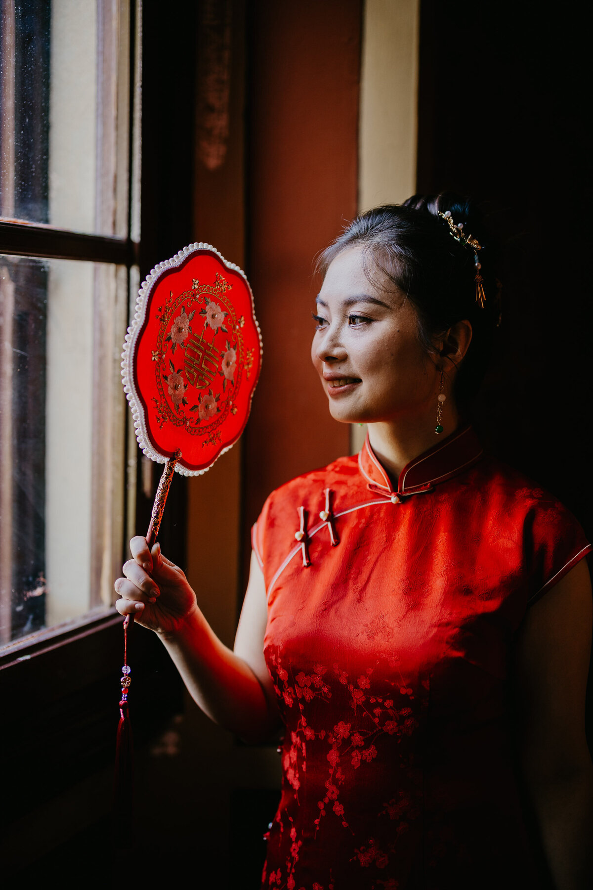 Bride in red Chinese dress holding fan by window, wedding photographer Tuscany.