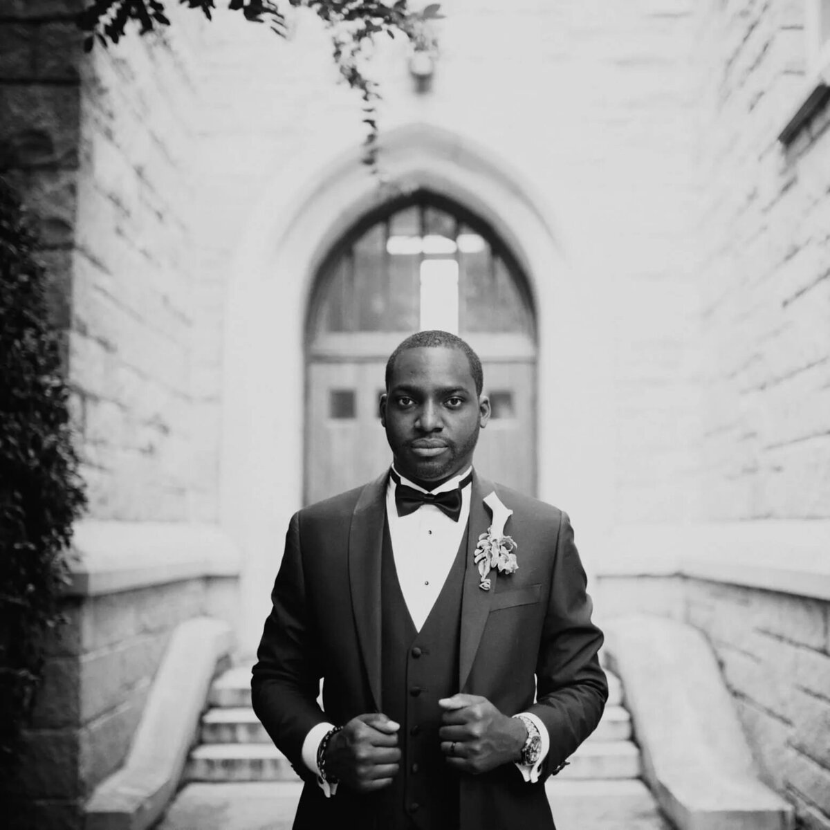 A groom in a black tuxedo stands confidently outside a stone building, his expression serious and poised.