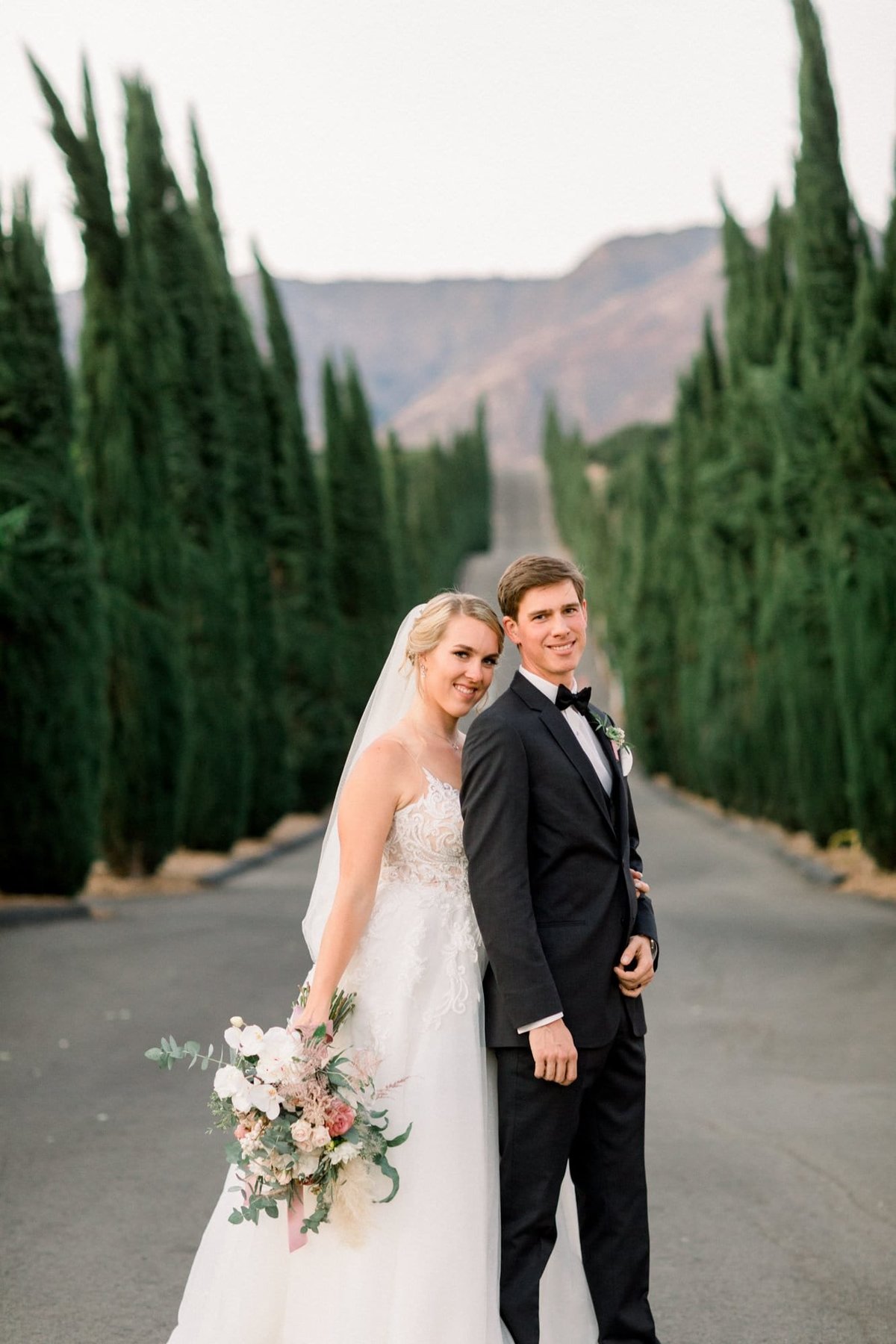 Bride and Groom pose together on a road between many tall bushes