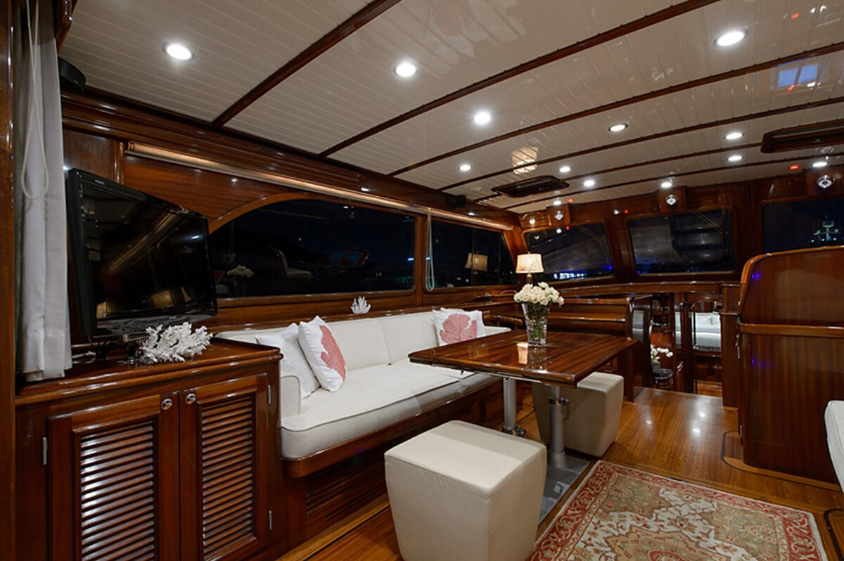 The interior of a luxurious yacht, featuring a polished wood finish, recessed lighting, and white leather seating, wooden table with a vase of white flowers is flanked by stools