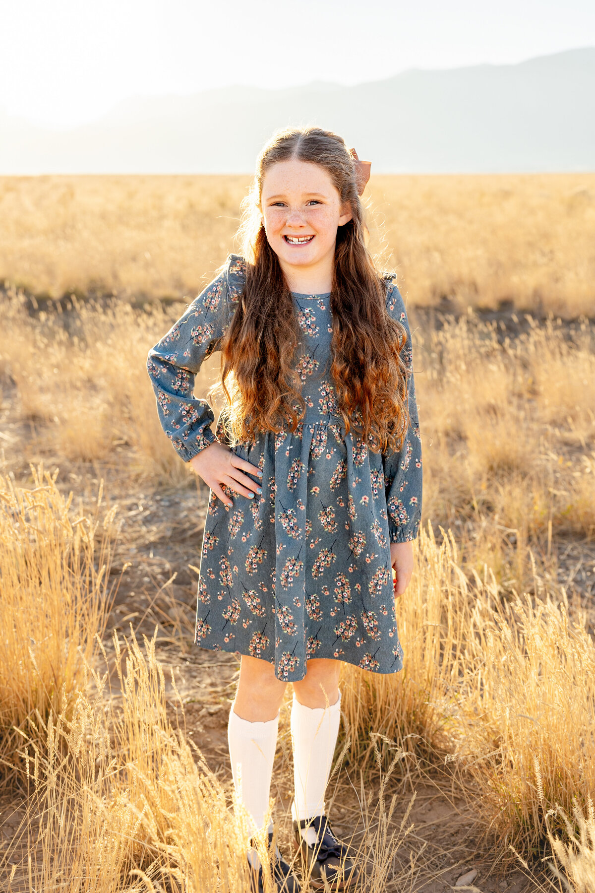 A young girl with long, wavy hair wears a blue floral dress while standing in a wheat-colored field in Eagle Mountain, Utah, during a family photoshoot.