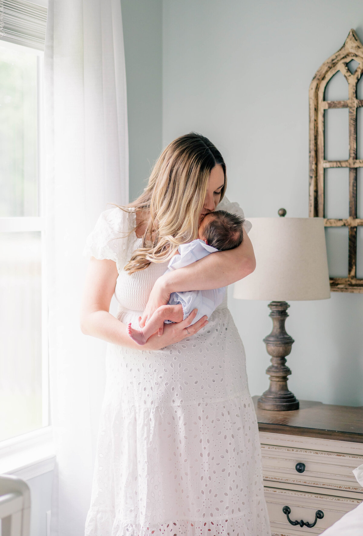 loving image of momma kissing her newborn child while standing in the sunlight from window during their indoor newborn session