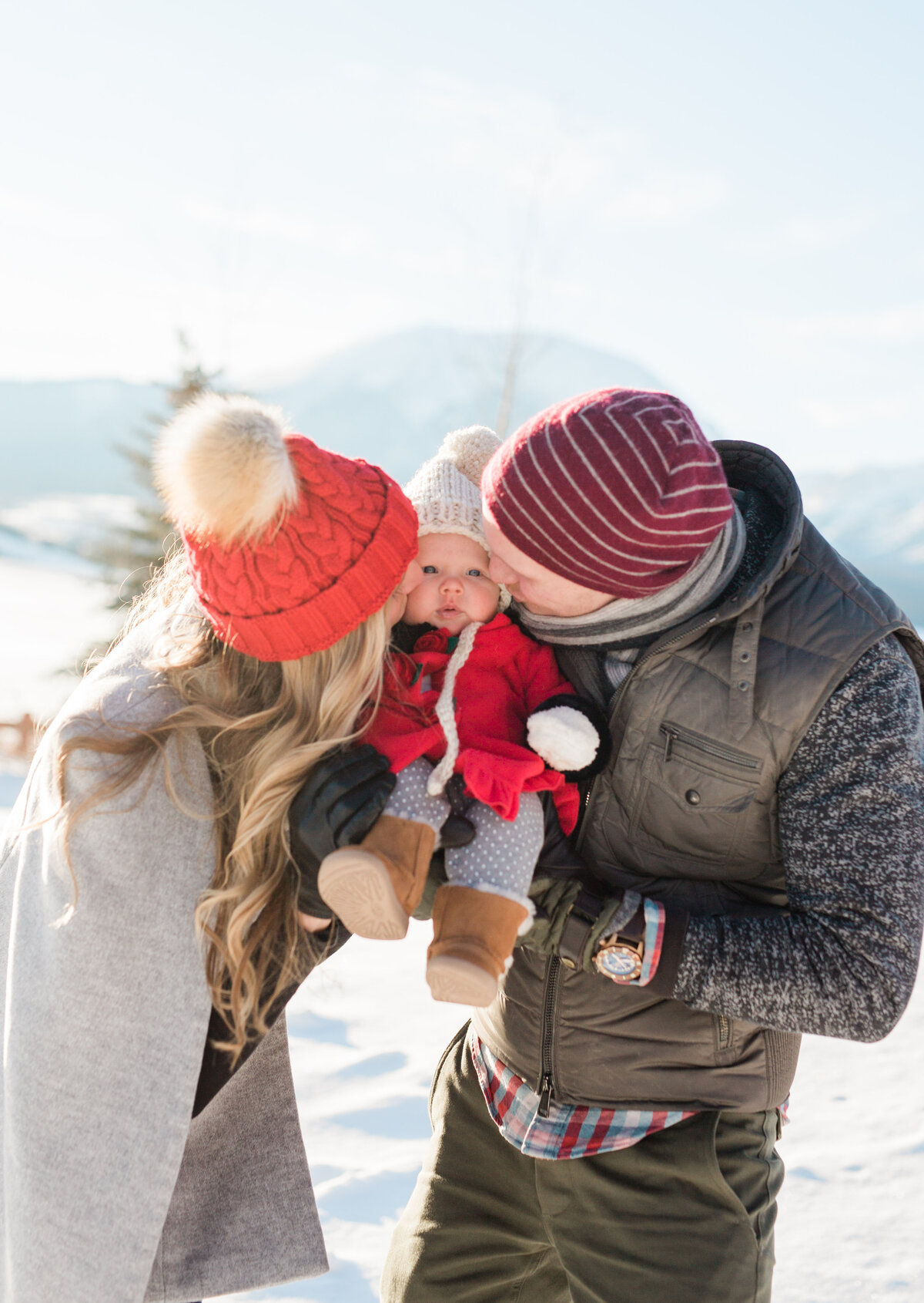 This family of 3 is tightly bundled up for winter. They have their newborn son between them are kissing him on each cheeck.