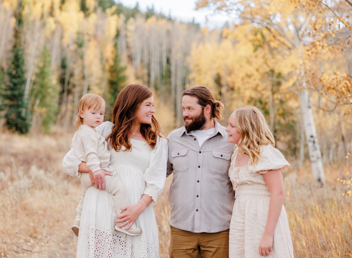 A mom and dad smile at each other while on a fall hike with teen daughter and baby
