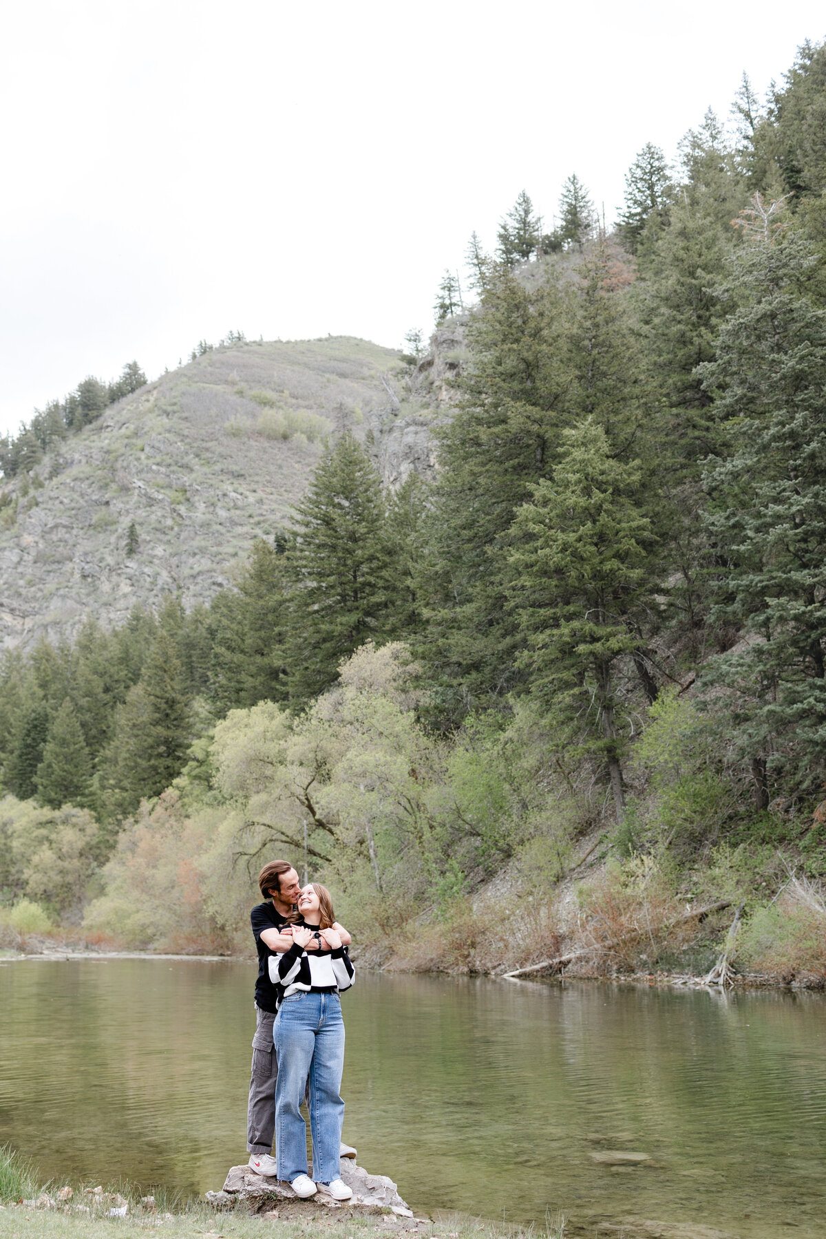 A couple leans into each other while standing on a rock near the river, framed by towering pines and rugged mountain scenery in Vivian Park, Provo Canyon.