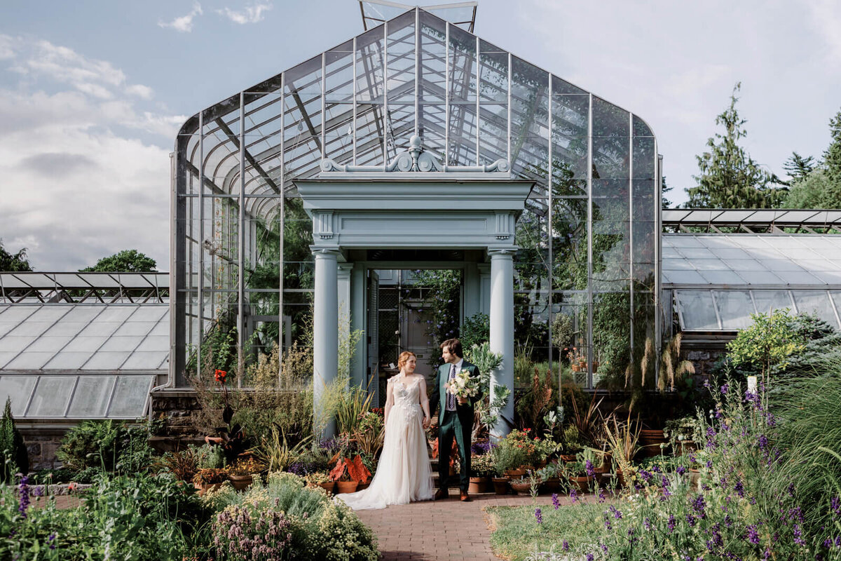 The bride and groom are standing in front of a greenhouse  with many plants around them