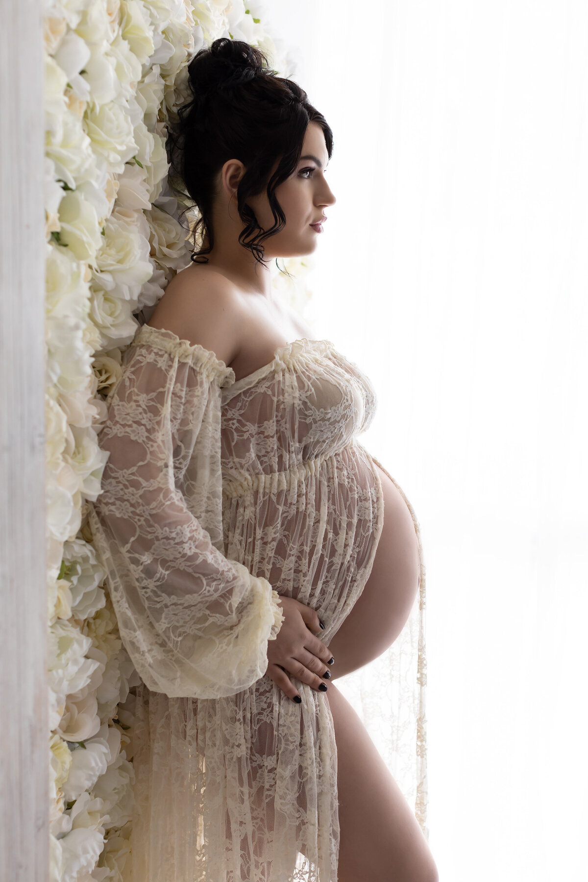 Profile view of a pregnant woman gently leaning against a wall adorned with soft white roses, wearing a delicate lace gown that enhances her graceful form. The bright natural light pouring in from the side creates a dreamy, airy effect, highlighting her serene expression. 