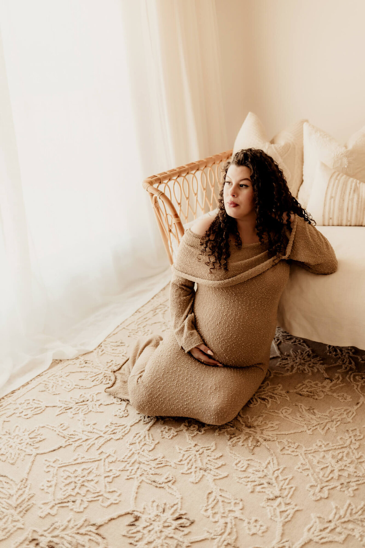 Woman with curly hair sitting on a rug next to a rattan boho daybed, Edmond maternity session.