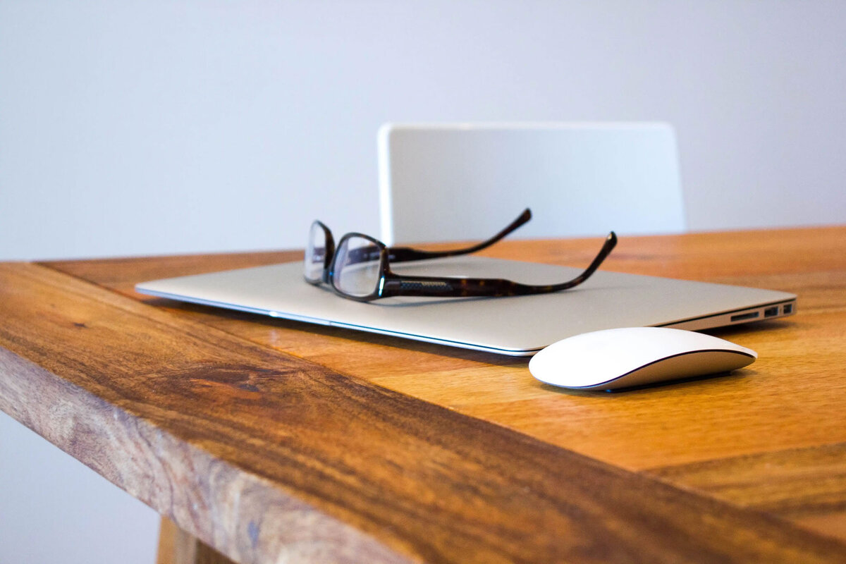 image of a silver laptop and white mouse sitting on a acacia table with a pair or reading glasses on top, all in front of a white wall