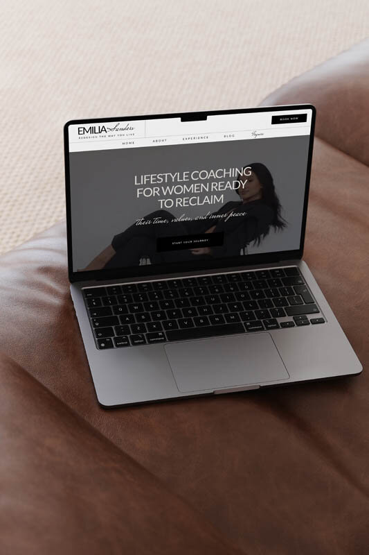 Laptop displaying Emilia Sanders Showit website homepage with bold lifestyle coaching headline, resting on brown leather surface