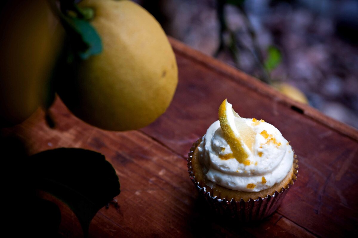 A lemon cupcake with fresh lemon shot on a wooden fruit box with lemons hanging from a tree in the foreground.