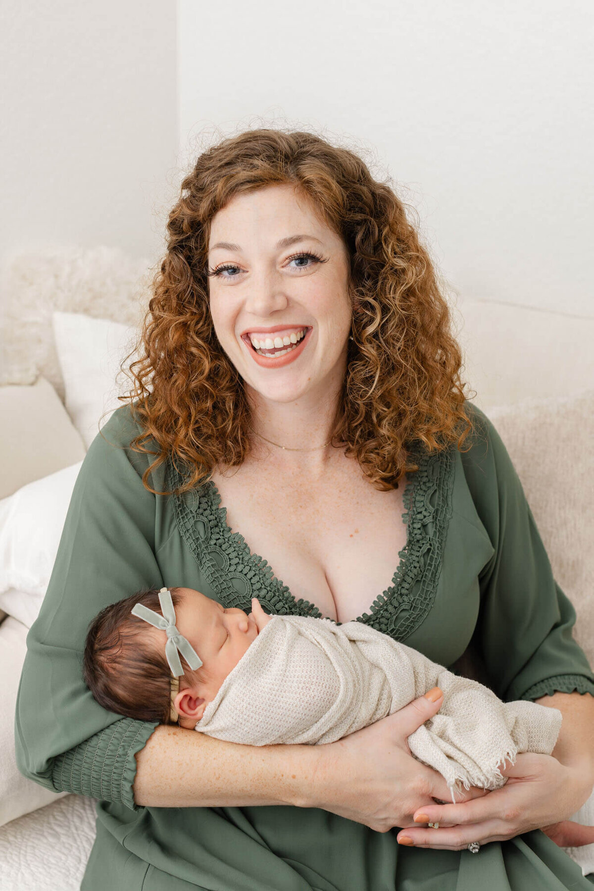 Mom in green dress, sitting down on bed while holding newborn baby girl. Baby is wrapped in a beige wrap with a velvet green bow on her head.