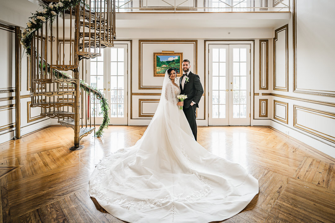 Bride and groom pose together in a grand mansion with spiral staircase and sweeping wedding gown train.