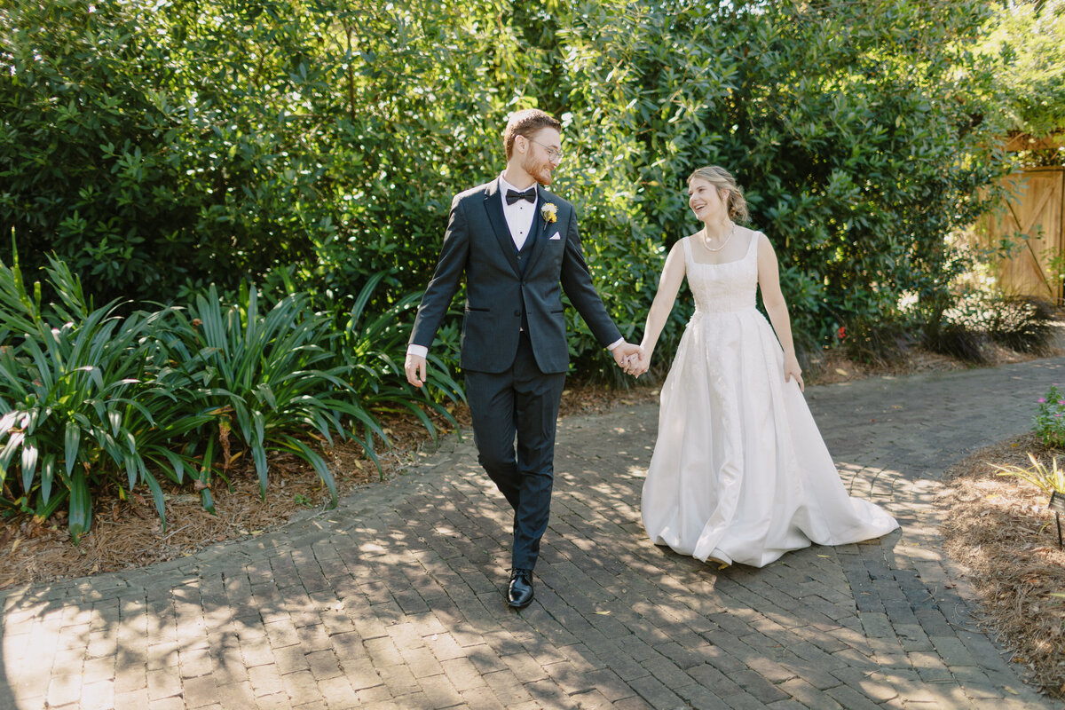 Red headed groom with glasses wearing black tux and bowtie looks over shoulder at his bride wearing a true white bridal gown and pearls as he leads her down cobble stone walkway in the. midst of lush greenery.