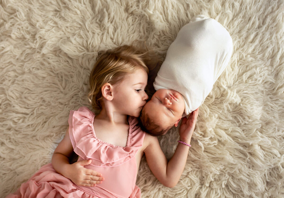 A newborn baby boy sleeps peacefully as his big sister gives him a  kiss on the cheek