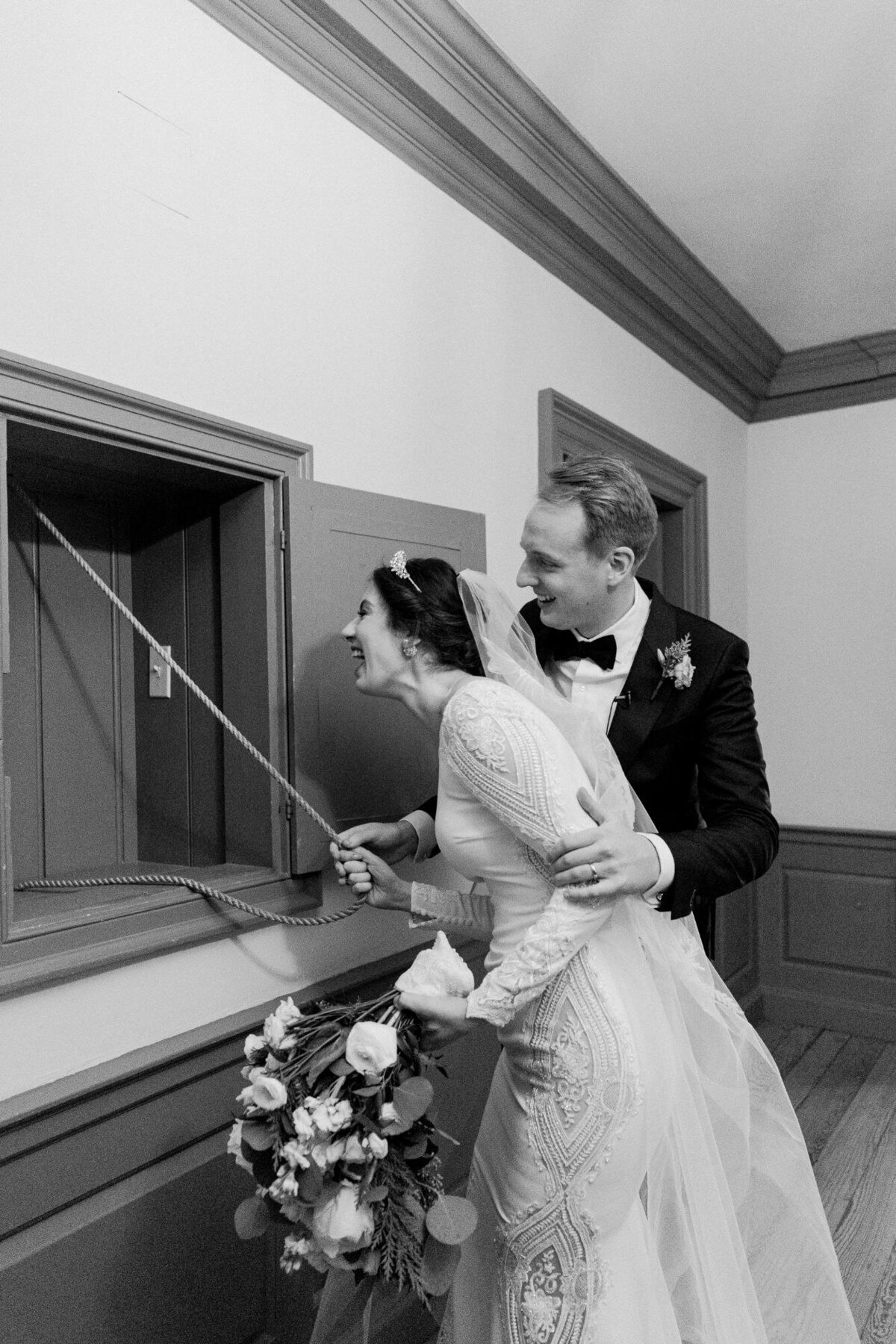 Bride and groom playfully tug on a window blind cord together in a black and white photograph.
