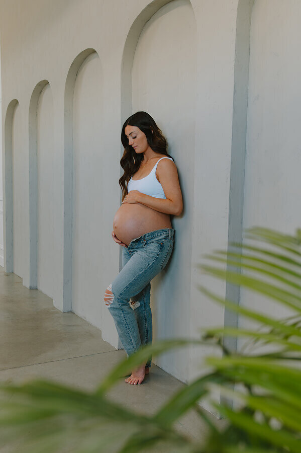 A pregnant woman poses for a candid portrait. She's leaned up against a wall and holding her belly.