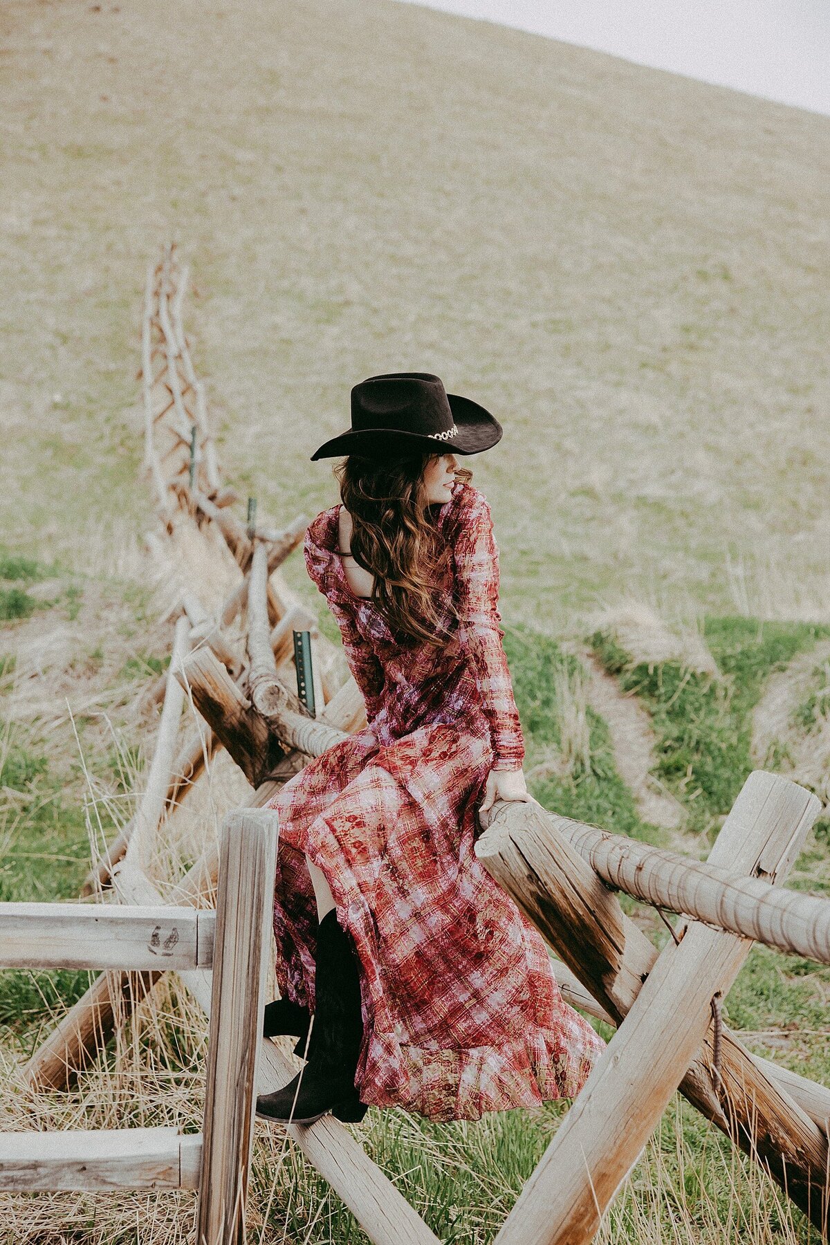 Senior girl in a long red and black patterned dress and black cowboy hat, perched on a rustic wooden fence in a rural field setting in Salt Lake City Utah