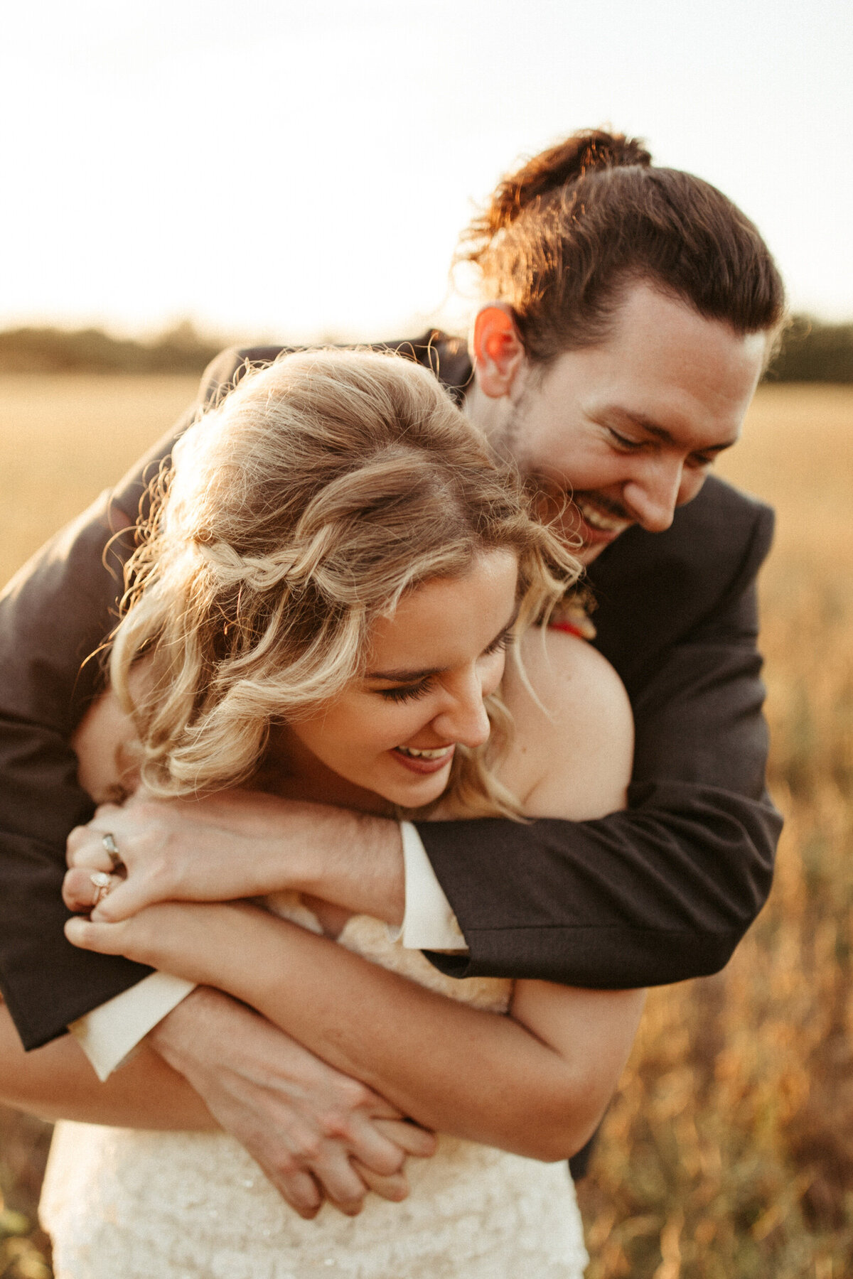 Groom wrapping his arms around his bride in a field at sunset as they laugh together