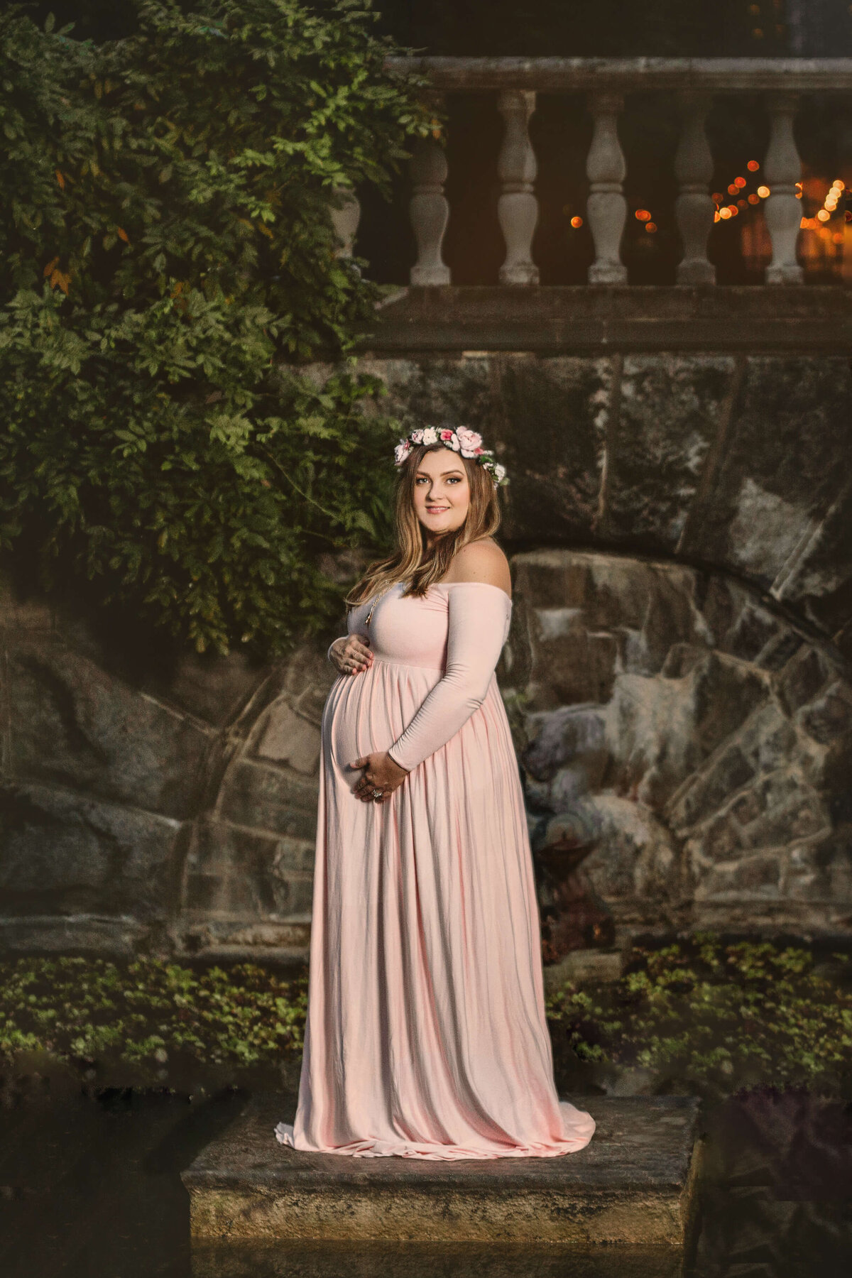 pregnant mom wearing a  pink maternity gown standing on a stone platform in front of a pond with lights in the background