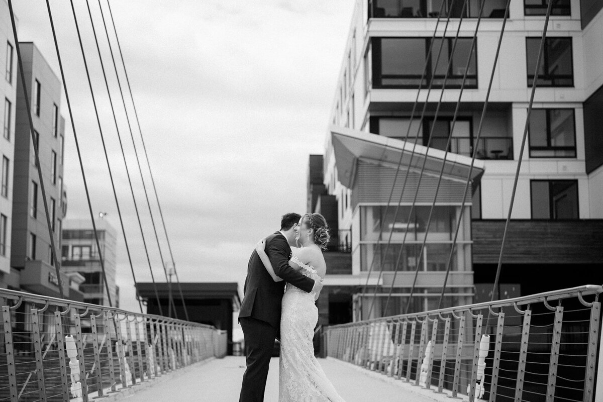 Bride and groom kiss under open skies with bridge in the background on beach in Boston, Massachusetts. 