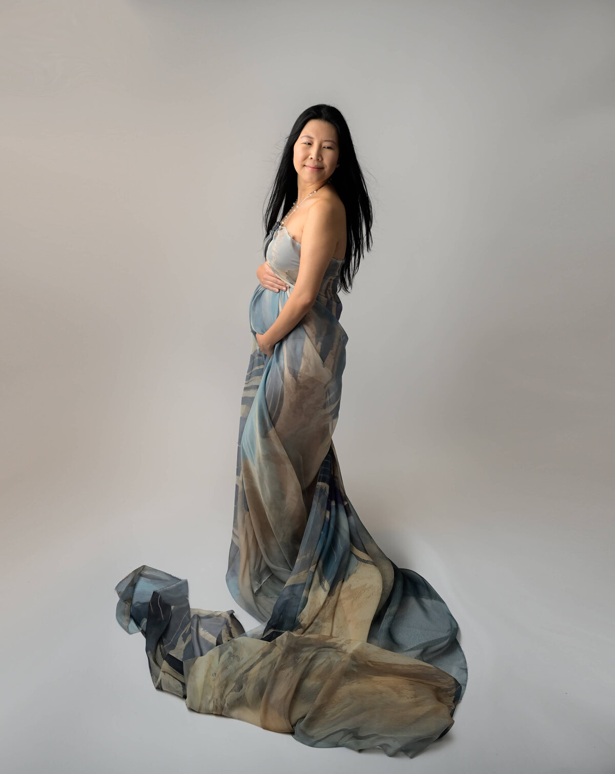 Mother-to-be in a side profile, looking over her shoulder, with flowing fabric tossed by a fan. Image captured by E. Benecki Photography.