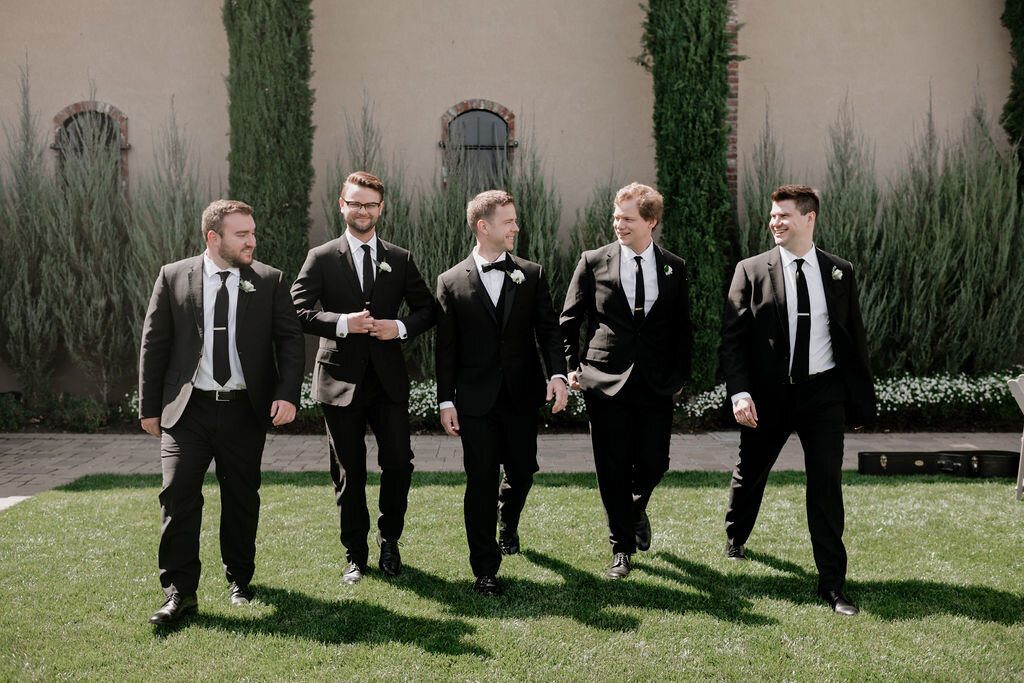 Groom and groomsmen walking and laughing in black tuxedos during a spring wedding at the Palm Event Center in Pleasanton, CA.
