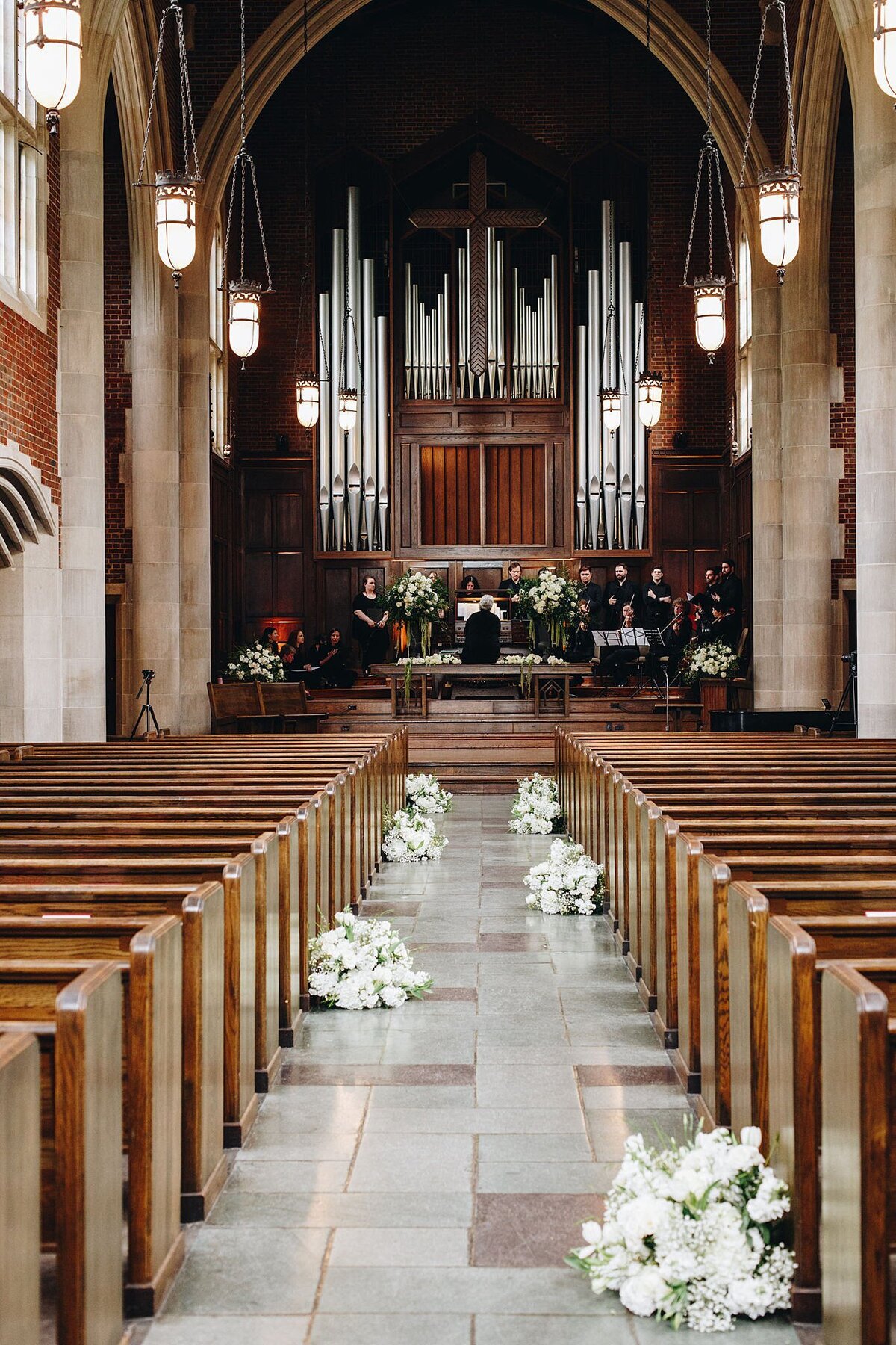 Church with many rows of brown wood pews lead up to the altar where an orchestra sits in front of a large pipe organ. The aisle is decorated with white floral arrangements.