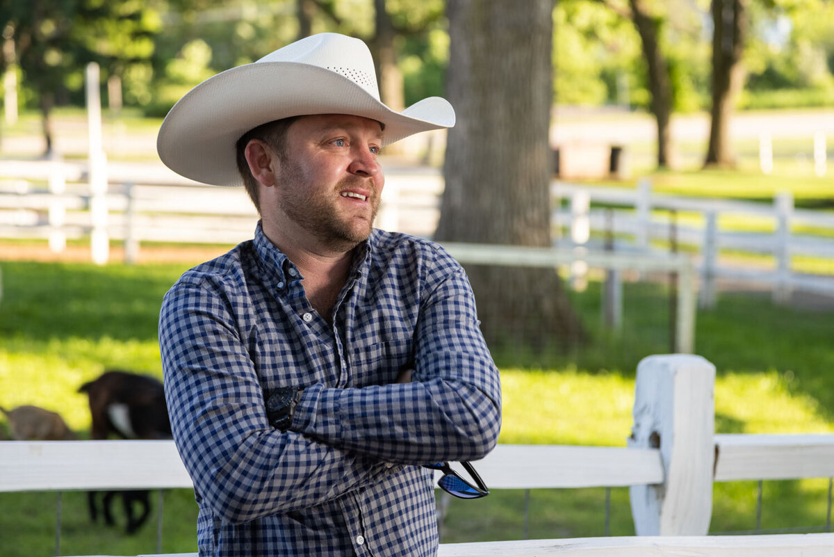 A man wearing a cowboy hat and plaid shirt is sitting on a bench with his arms crossed