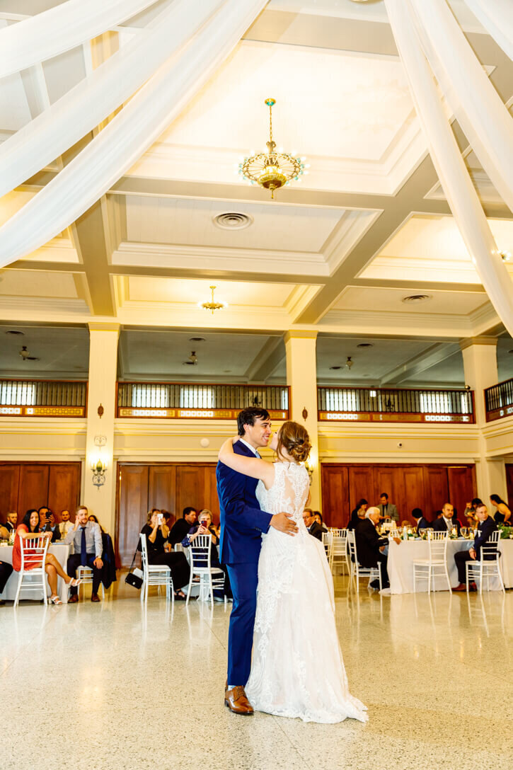 Bride and Groom sharing their first dance in the ballroom underneath a beautiful chandelier and their guests all watching.