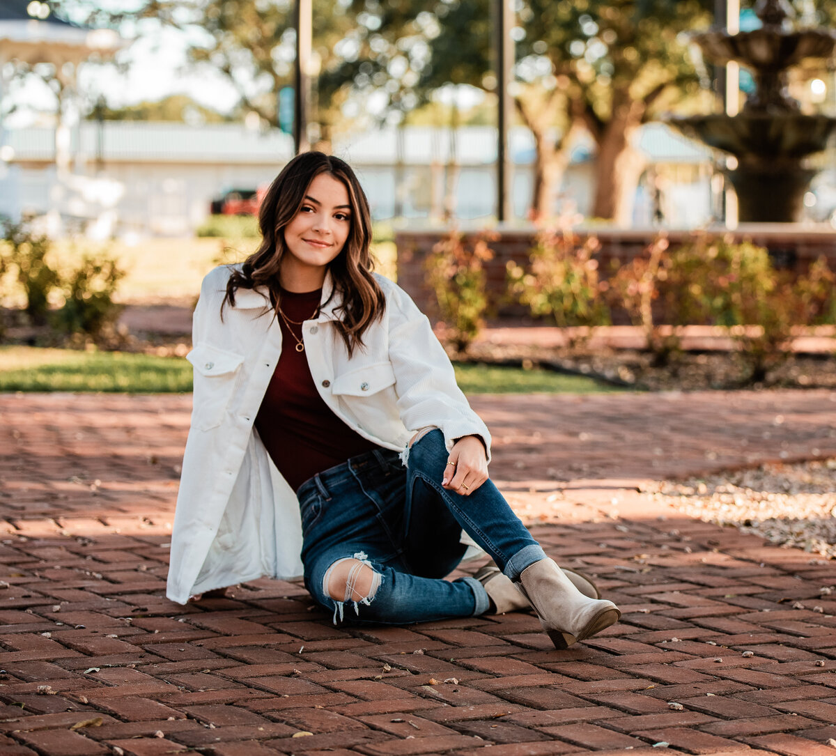 A senior girl sits on a red brick walkway for her graduation pictures.