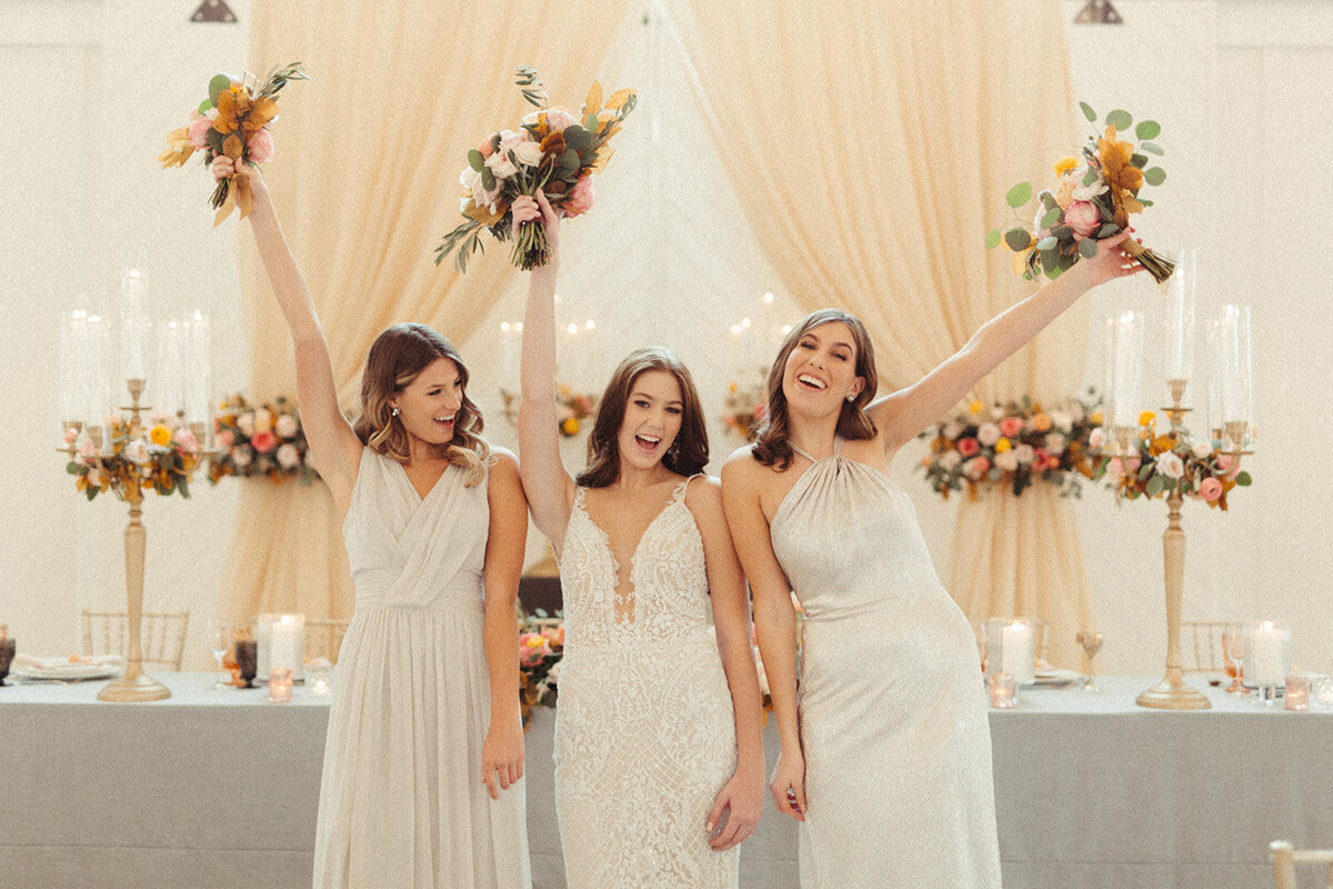 A bride wearing a white wedding gown poses with bridesmaids wearing light gray gowns holding bouquets in the air, smiling.