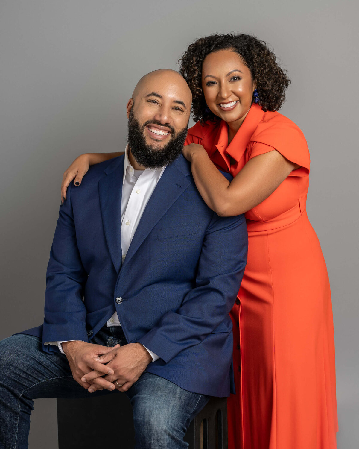 Couple posing, the man in a blue suit and the woman in an orange dress, both smiling.