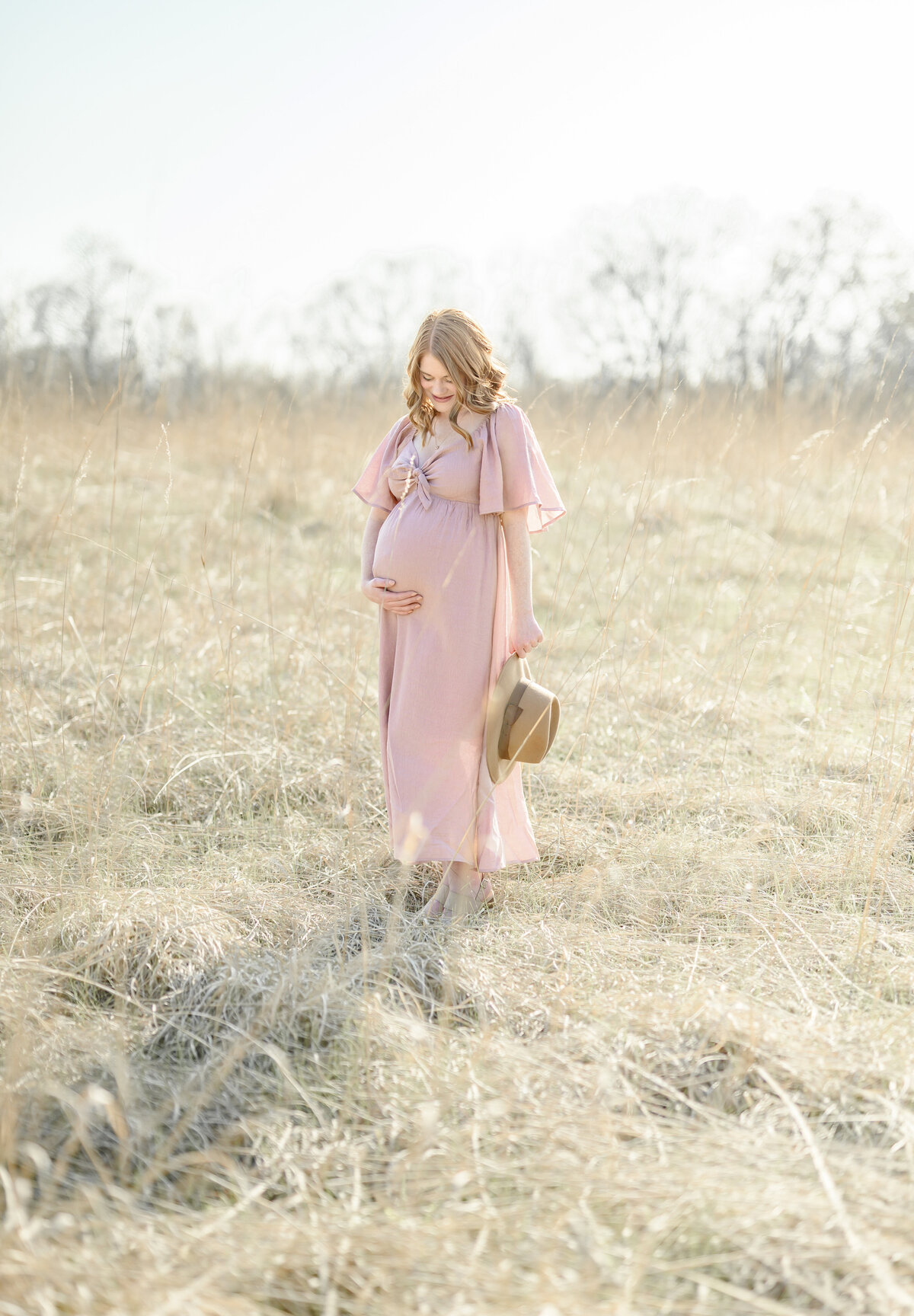 Expectant mother in a pink dress walking through a sunlit field with tall grass.