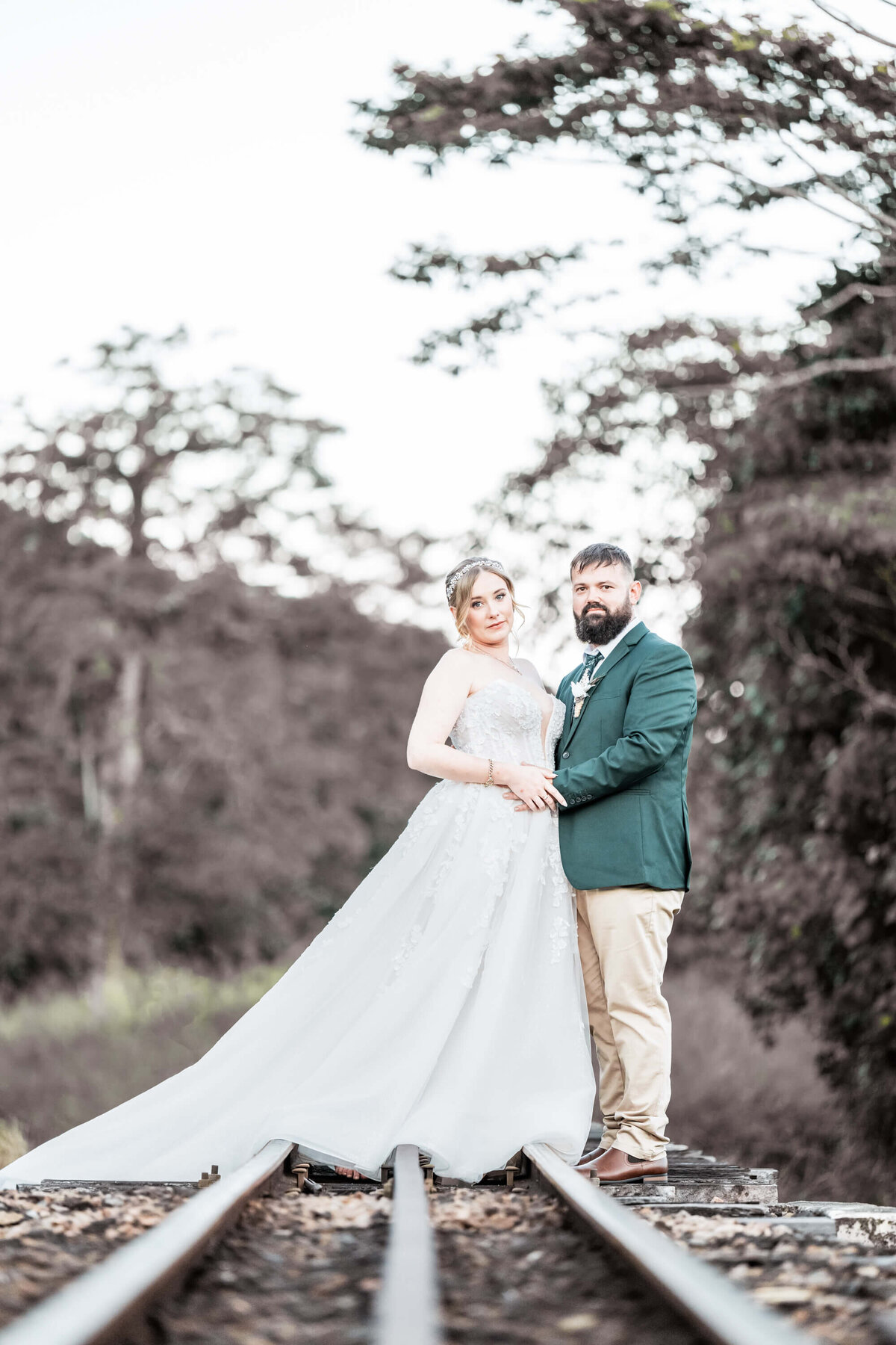 The bride and groom stand on rustic train tracks, enveloped by trees, embodying romance and the journey of marriage. - photo by Jamie Simmons