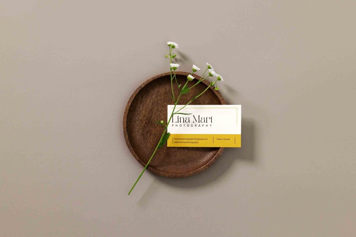 Lina Mart Photography business card with yellow accent placed on a wooden dish beside a sprig of white flowers