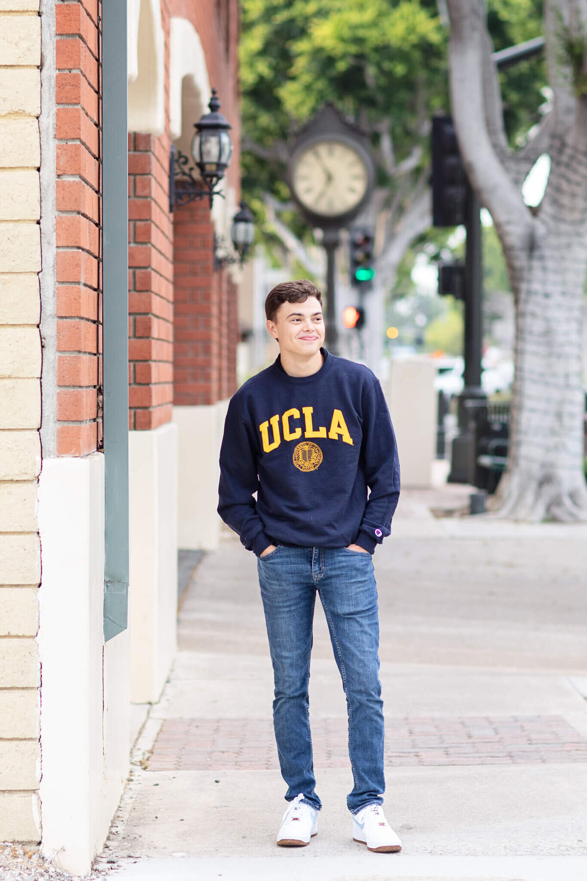 A person wearing a navy UCLA sweatshirt stands casually on a sidewalk near brick buildings. In the background, theres a clock and greenery. The person is smiling and looking to the side.