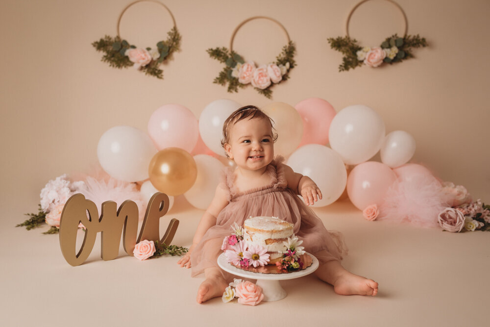 Photo shoot with Atlanta, Georgia cake smash photographer with one year milestone cake smash at Marietta GA portrait studio with baby girl wearing pink tulle dress sitting on floor eating cake decorated with flowers. Balloons are behind her with a gold O N E sign and floral decor on the tan backdrop