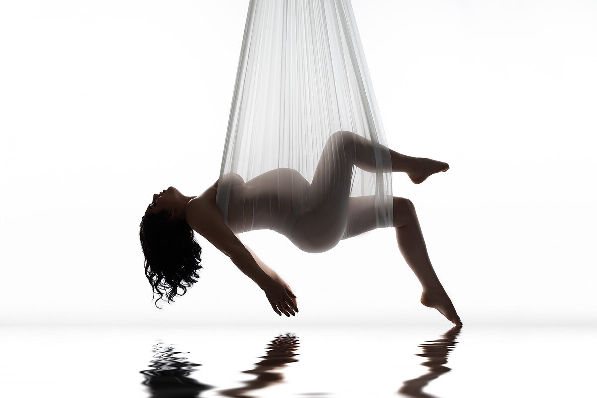 DFW maternity photography of woman gracefully suspended in white fabric above water reflection.