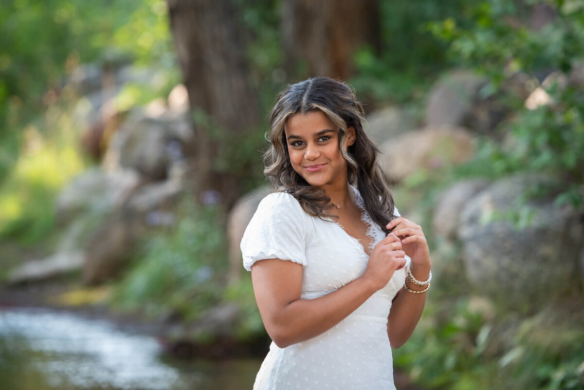 A woman stands in a creek wearing a white dress while twirling her hair