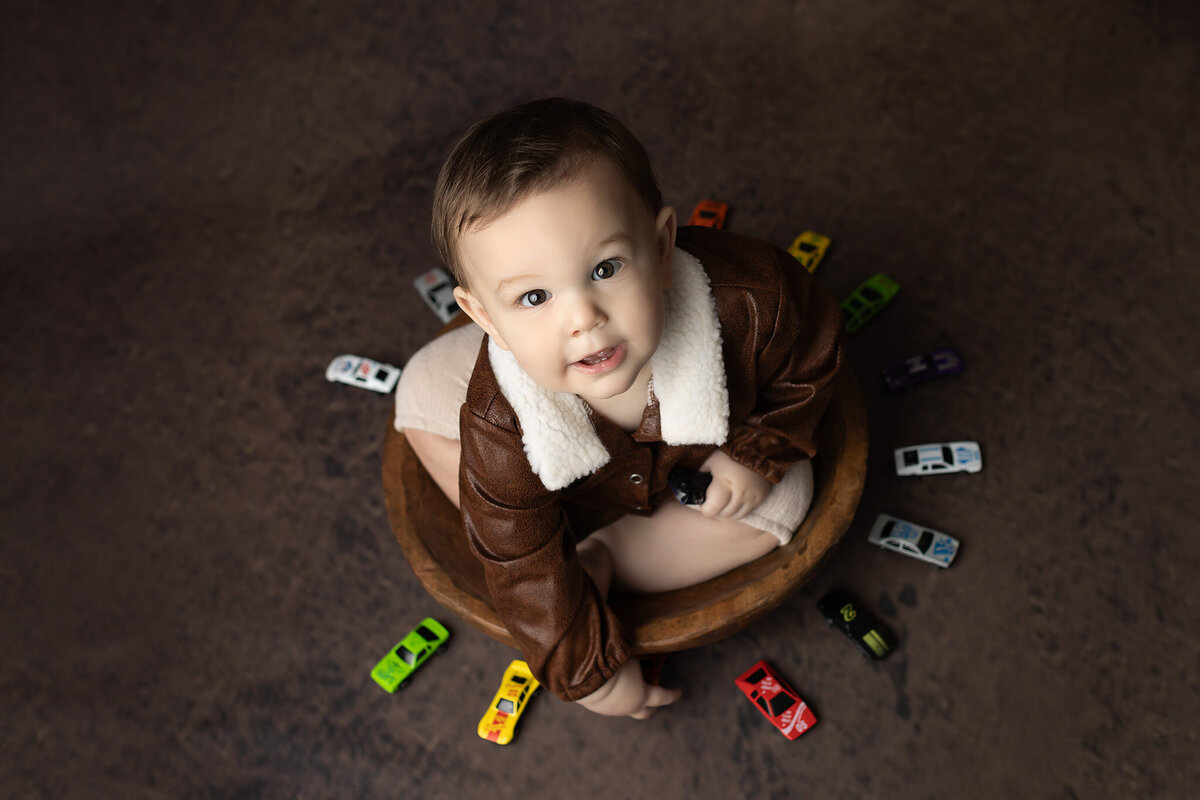 Little boy dressed in a brown aviator jacket sitting in a wooden bowl on a dark studio floor, surrounded by colorful toy race cars, captured by a Dallas milestone photographer.